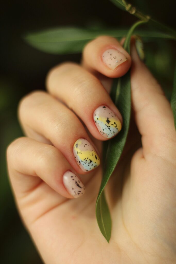 Short round nails with abstract speckled nail art &mdash; sheer base with black flecks and a yellow color-block stripe, hand holding green leaves &mdash; abstract nail color trends 2026