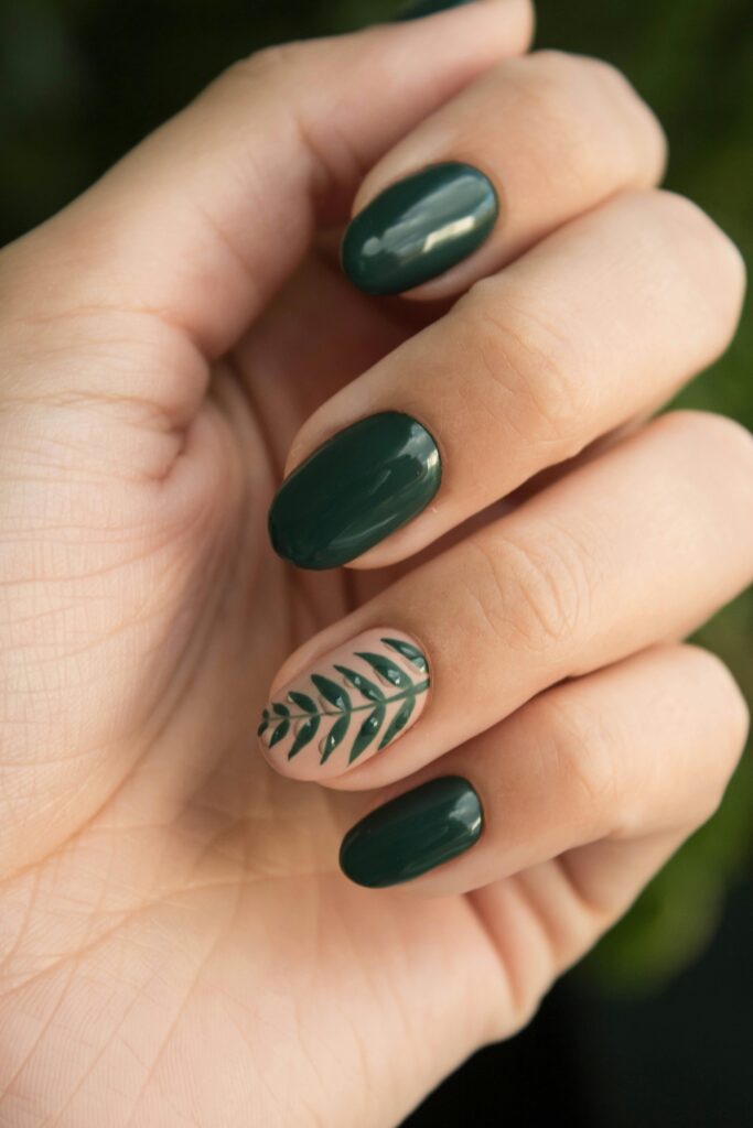 Hands with dark grey-green nail polish wearing gold stacked rings, resting on a bed of glossy green leaves &mdash; moody nail color trend 2026