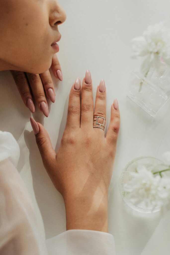 Woman with long almond nude-pink nails wearing a silver geometric ring, hands resting against a white surface with white flowers &mdash; warm nude nail color trend 2026