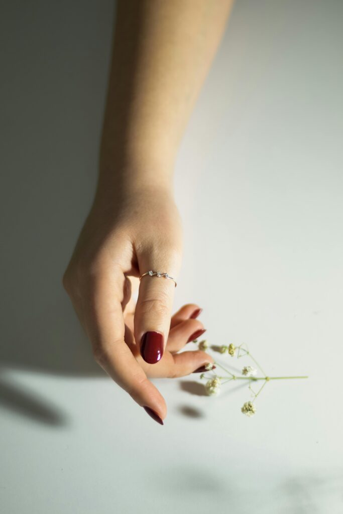 Close-up of a woman's hand with deep burgundy red nails wearing a delicate crystal ring, reaching toward dried flowers on a white surface &mdash; dark red nail trend 2026