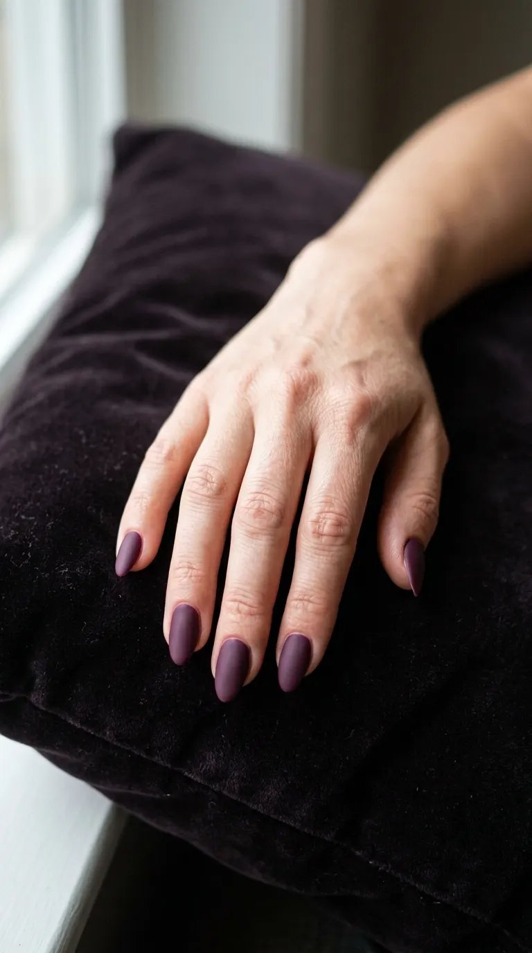 Matte plum nails on a hand resting on a dark plum velvet cushion