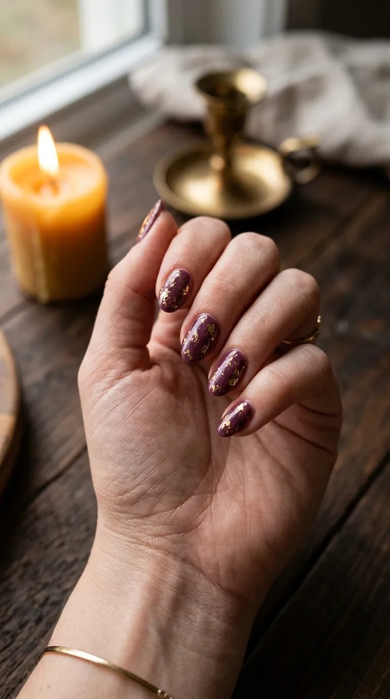 Plum nails with gold foil flakes beside a lit candle on dark wood